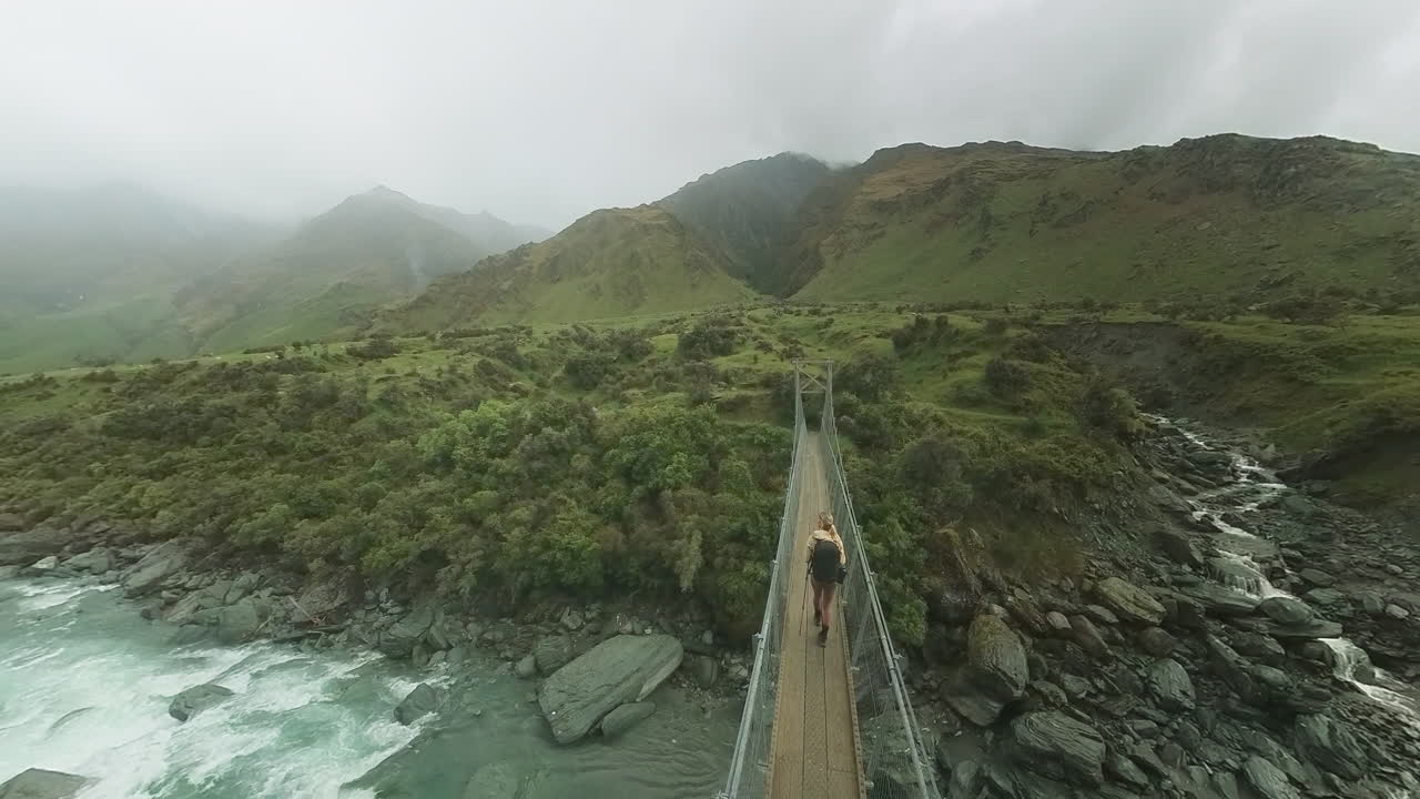 mujer cruzando un pequeño puente colgante en el remoto valle lluvioso de nueva zelanda