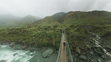 woman crossing small suspension bridge in remote rainy valley new zealand