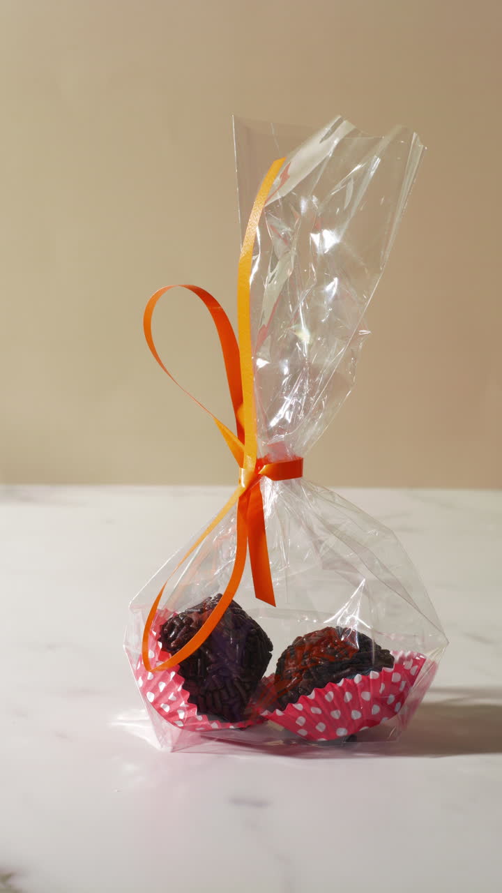 Close-up of a hand taking a clear gift bag containing two chocolate truffles in red polka dot wrappers, tied with an orange ribbon. Bright lighting highlights the festive and elegant presentation.
