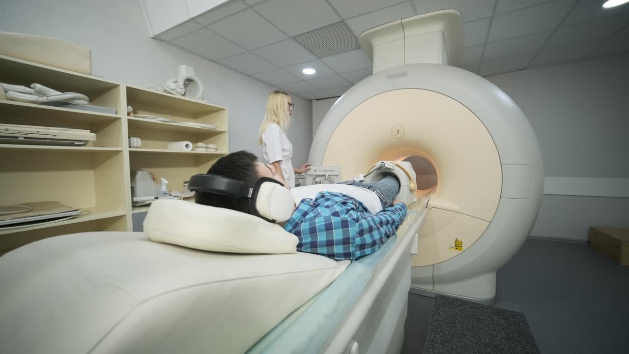 여성 의사는 무 관절 mri 스캔을합니다. 자동 테이블에 있는 젊은 남성 환자는 소음 격리 헤드폰을 사용하여 폐쇄형 mri 기계에 들어갑니다. 현대 장비, 환자의 무에 코일
