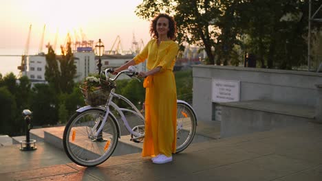 Lächelnde-Brünette-Frau-In-Einem-Langen-Gelben-Kleid-Steht-Neben-Ihrem-Stadtfahrrad-Und-Hält-Den-Lenker-Mit-Blumen-Im-Korb