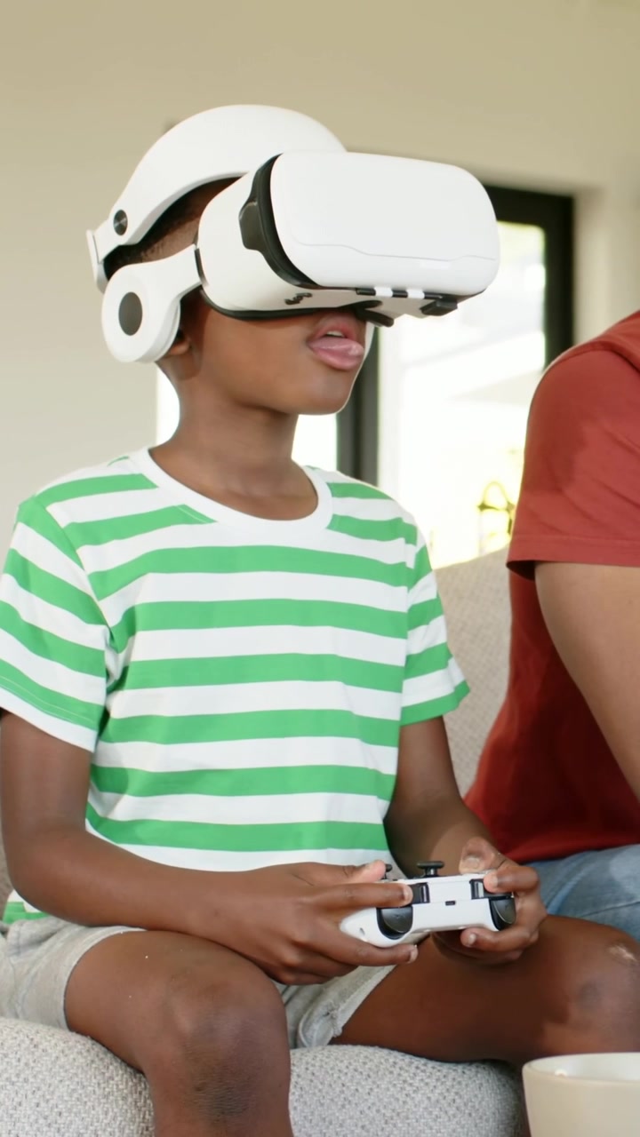 Video verticale: Bambino/a in maglietta a righe reagisce premendo i pulsanti di un controller bianco a casa mentre la VR inizia.