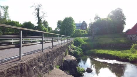 Conducción-De-Automóviles-En-Un-Viejo-Puente-De-Piedra-Junto-A-Un-Arroyo-Con-Casas-En-El-Fondo,-En-El-Sur-De-Suecia-Skåne-Österlen-Svenstorp-Antena-Baja-Adelante-Lento