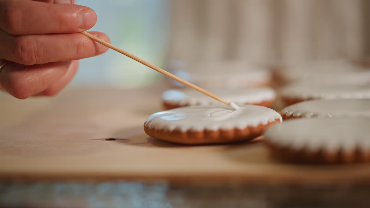 Closeup hand icing scalloped cookie on wooden board, home baker gently spreads smooth white glaze with toothpick, rows of iced cookies cooling in soft natural light, warm cozy kitchen atmosphere
