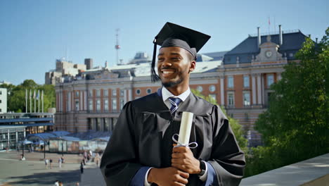 happy young man in graduation cap and gown holding his diploma on a university campus