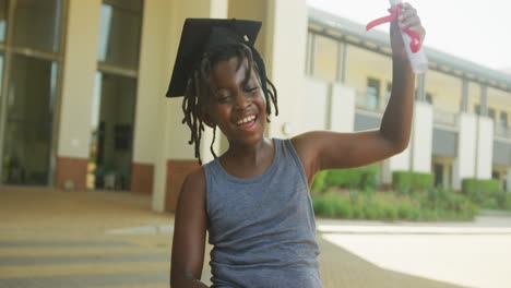 Video-of-happy-african-american-boy-wearing-graduation-hat-and-holding-diploma-in-front-of-school