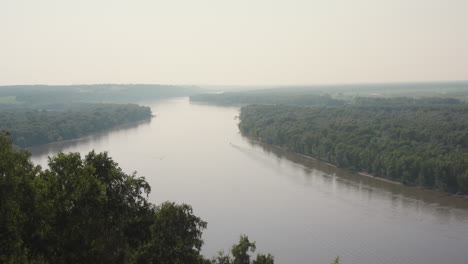 peaceful countryside view featuring wide winding river bordered by dense green forest beneath hazy morning sky, radiating calm and tranquility across rural landscape