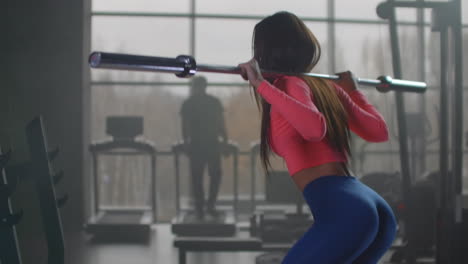 la joven con la camiseta rosa y las leggins azules se está preparando para competiciones importantes. está haciendo cuclillas con una barra vacía con una técnica correcta en el gimnasio en el entrenamiento de la mañana.