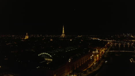 Night-fly-above-illuminated-Seine-river-waterfront.-Les-Invalides-and-Eiffel-tower-landmarks-above-town-development.-Paris,-France