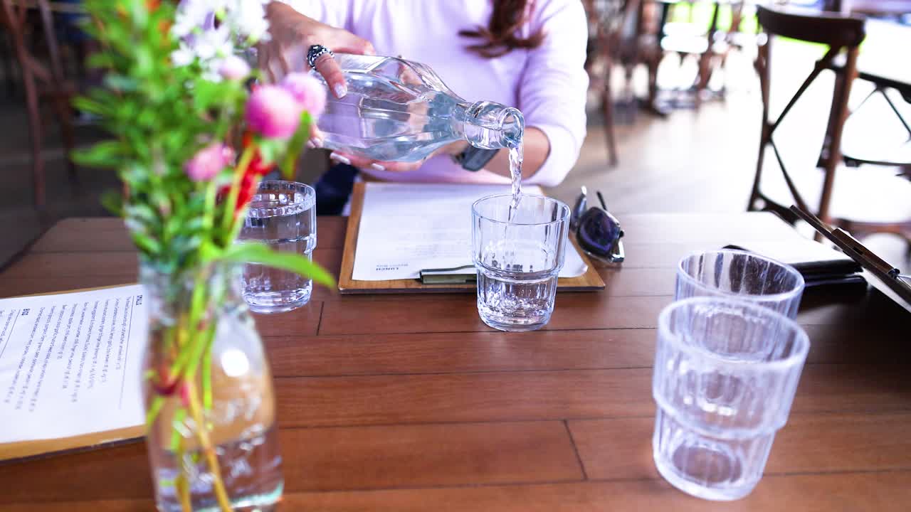 A person pours water into glasses at a sunlit cafe table, surrounded by flowers and documents