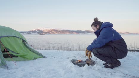 man in winter clothing sitting by the fire outside camping tent in snow at daytime