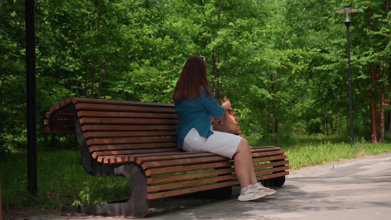 Young woman in white outfit and blue shirt walking along sunny park pathway toward wooden bench surrounded by lush green trees, enjoying peaceful summer atmosphere and gentle breeze