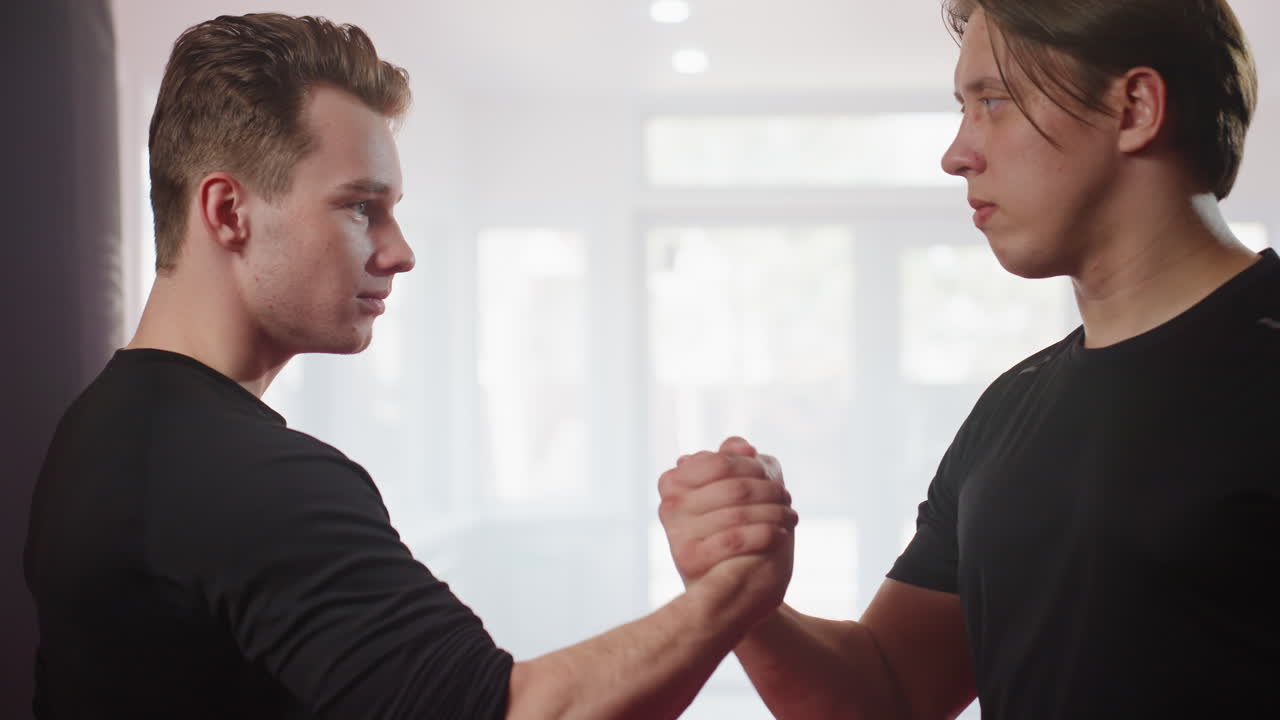 Two fighters in black sportswear face each other inside bright gym, standing firm with focused expressions, as they shake hands embodying confrontation and mutual respect