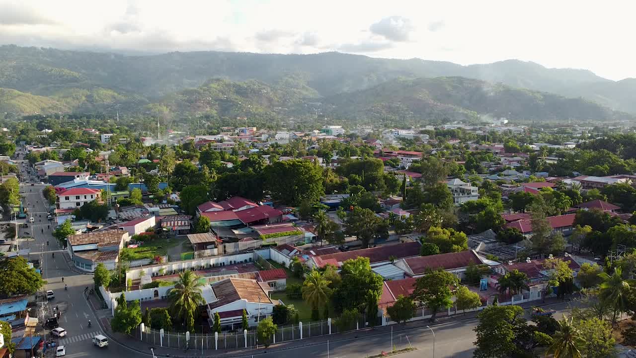 Aerial view lowering over capital city of Dili, Timor-Leste with red tinned roof houses, buildings, and inner city traffic with rugged mountains in distance