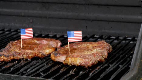 a wonderful shot of two juicy rib-eye steaks sitting on the grill and cooking with two small american flags on top of them with toothpicks