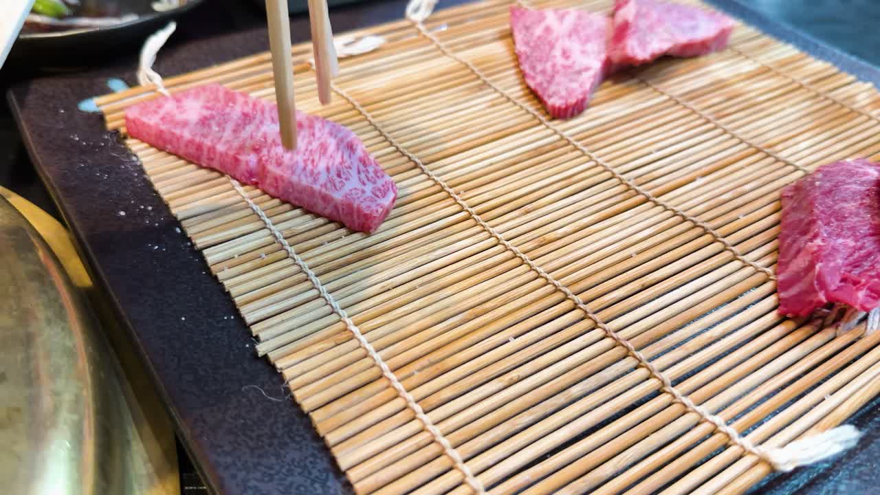 Primo piano di bacchette che afferrano carne di manzo cruda marmorizzata da una stuoia di bambù.