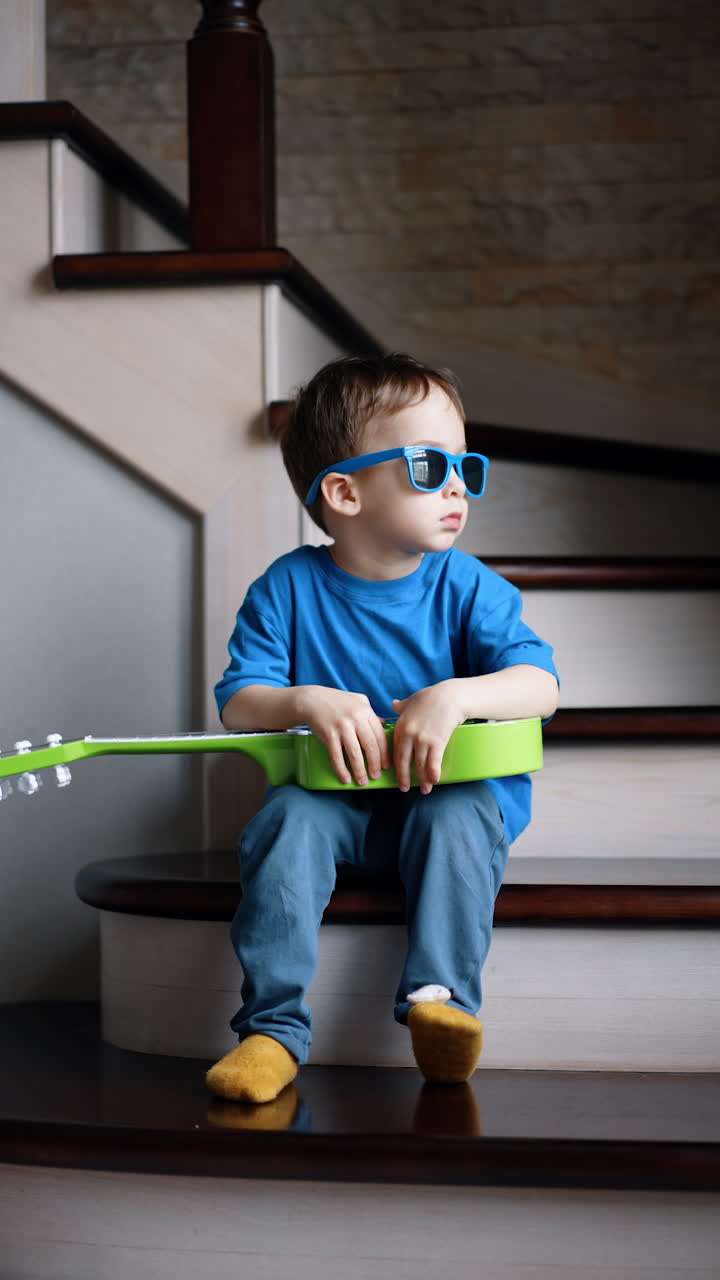 Adorable toddler boy wearing blue t-shirt and pants, yellow socks and sunglasses sits on the stairs indoors. Kid holds a guitar thoughtfully looking aside and then starts to play the instrument. Vertical video.
