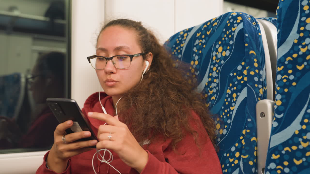 Commuter on train using smartphone, curly hair glasses red hoodie patterned seat window reflection evening light, student reviewing notes, traveler listening to podcast, remote worker replying