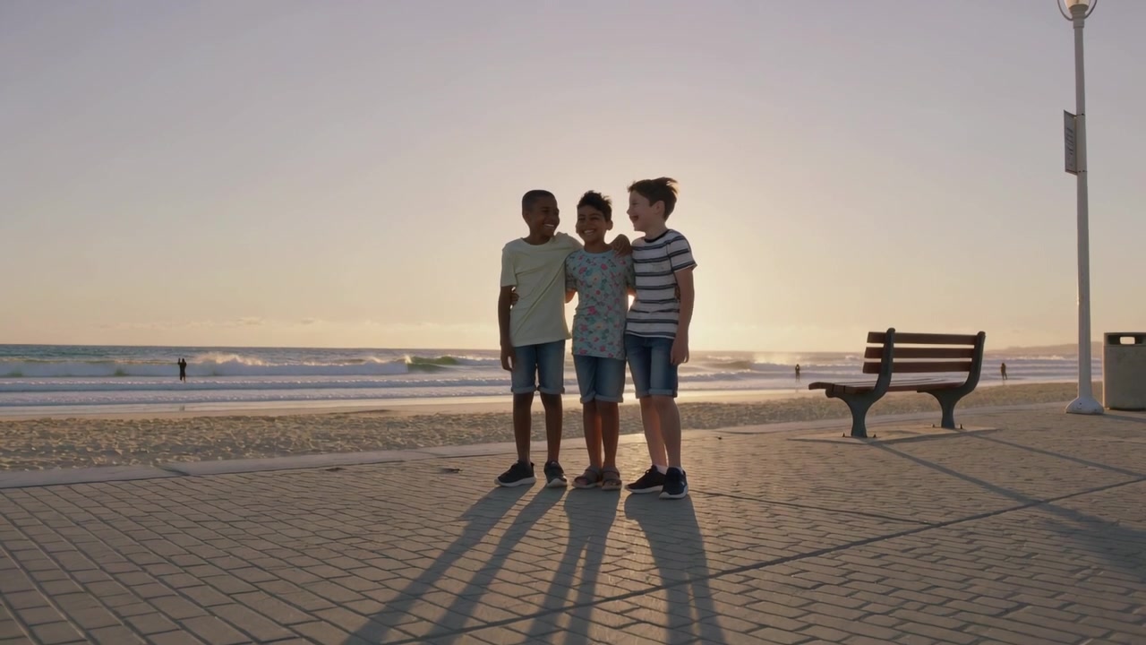 Tre ragazzi in piedi sul lungomare al tramonto, che muovono le braccia, si girano verso il sole, rimanendo vicini e indossando magliette.