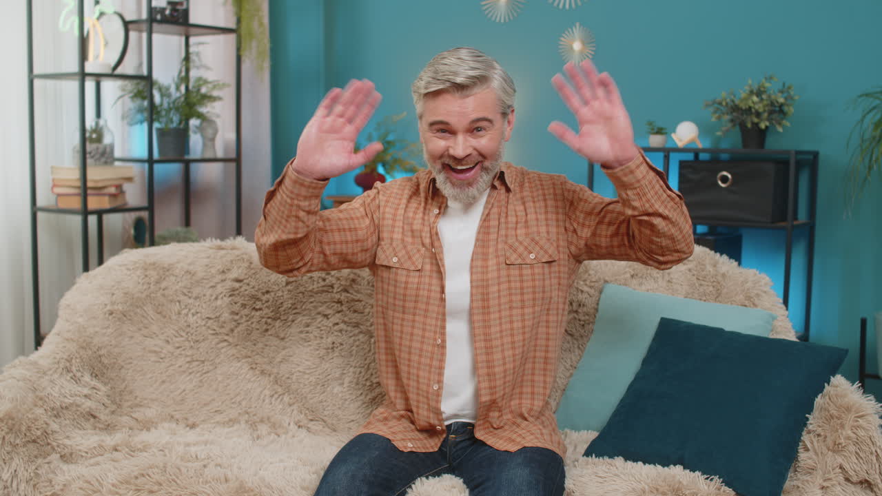 Mature man smiling friendly sitting on cozy sofa waving hand in greeting hello hi at home room