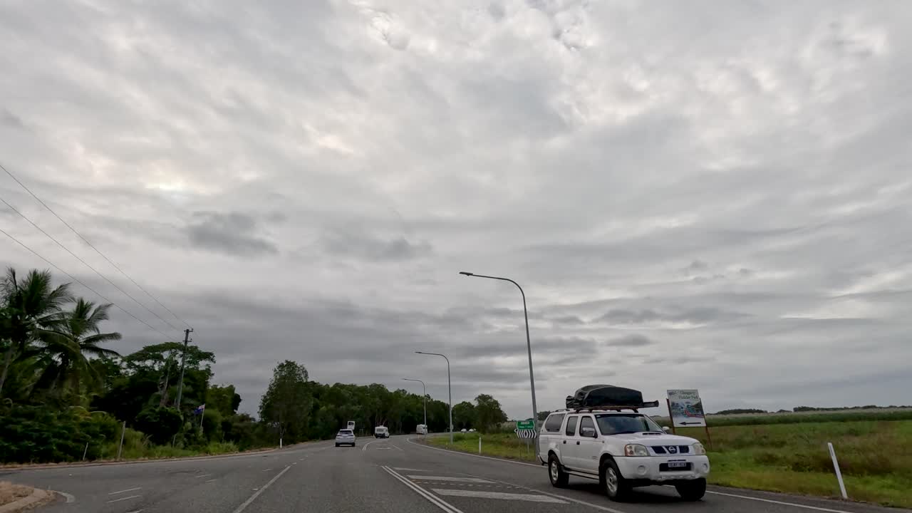 A drive along a highway in Port Douglas, Australia, showcasing overcast skies, sugarcane fields, and passing vehicles