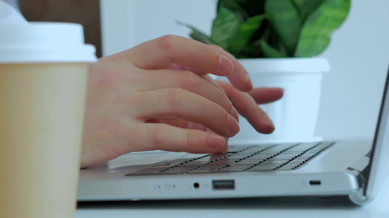 Male hands print the text on the laptop keyboard on the white table, a green indoor plant on the background, a paper disposable glass with coffee. Without a face, a close -up