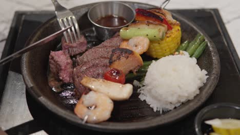 eating rare cooked beef steak with fork and steak knife, served with white rice and shrimp kebab in a sizzling plate