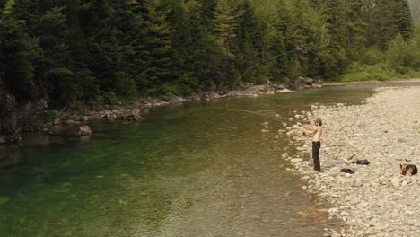 Hombre-Pescando-Con-Mosca-Con-Un-Perro-En-Un-Río-Poco-Profundo-En-Mcdonald-Creek,-Amplia-Toma-De-Seguimiento-De-Drones