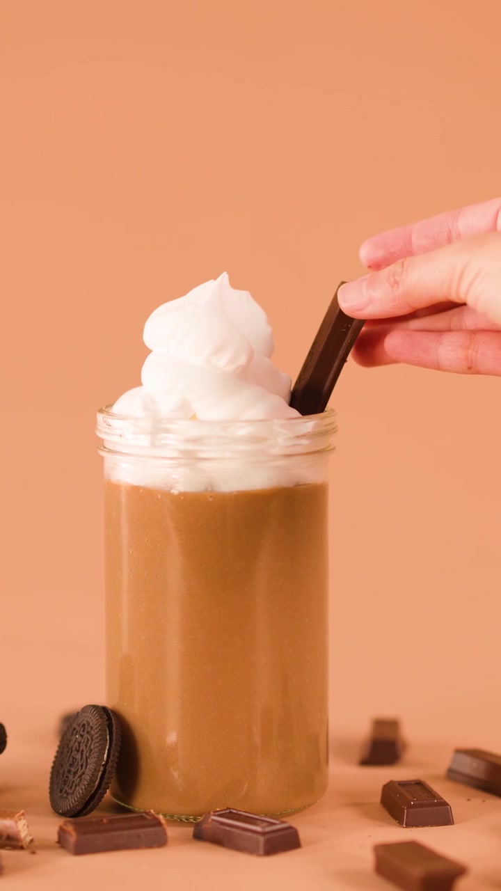 Una mano posiziona una barretta di cioccolato in un milkshake con panna montata, circondato da biscotti e pezzi di cioccolato