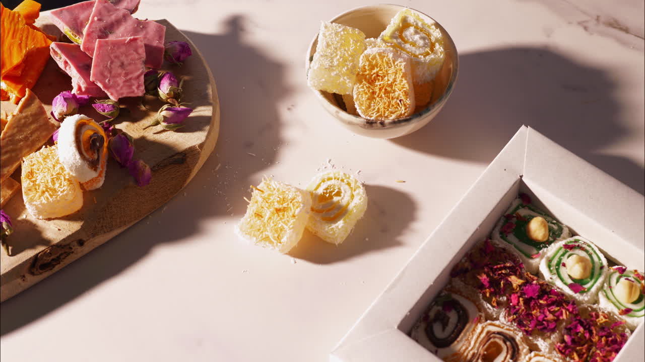 A hand reaches for a yellow swirled Turkish delight with kataifi topping beside a gift box of assorted lokum, wooden board featuring pink strawberry chocolate, brittle, dried rosebuds in warm sunlight