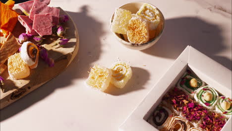 a hand reaches for a yellow swirled turkish delight with kataifi topping beside a gift box of assorted lokum, wooden board featuring pink strawberry chocolate, brittle, dried rosebuds in warm sunlight