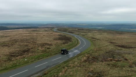 Drone-tracking-a-black-4x4-truck-driving-down-very-long-empty-straight-scenic-road-surrounded-by-hills-and-fields-of-moorland-in-the-North-Yorkshire-Moors-on-very-cloudy-overcast-day