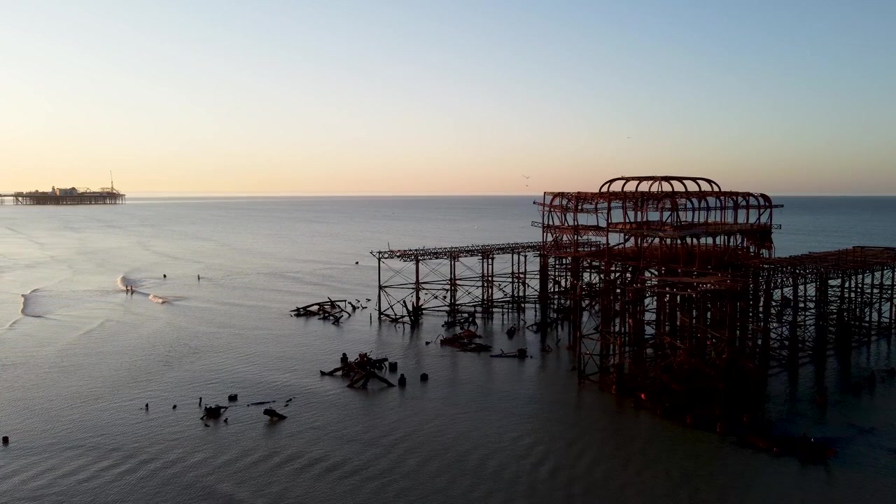 Drönarnärbild av ruinerna av Brighton West Pier med utsikt mot den östra piren när solen går upp över havet och stadssiluetten