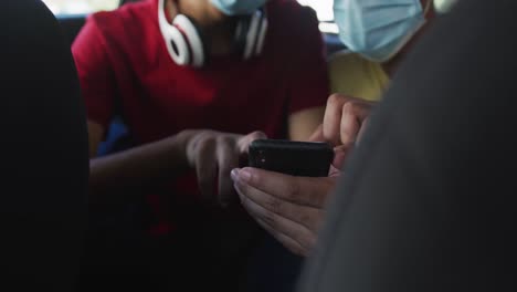 dos amigos varones de raza mixta que usan máscaras faciales usando un teléfono inteligente en un taxi