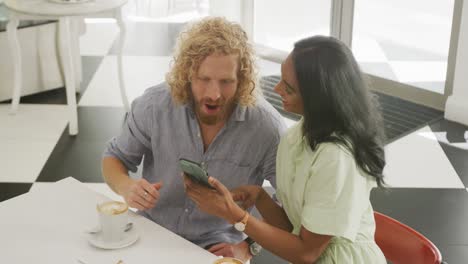 happy diverse couple with coffee using smartphone and talking at a table in cafe