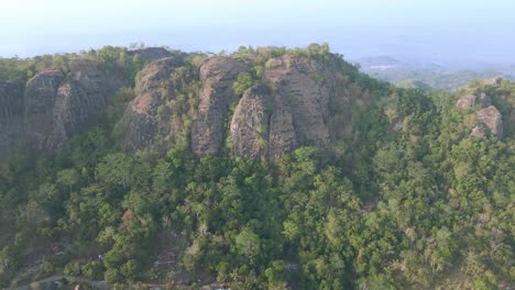 Sliding-drone-shot-of-huge-rock-mountain-surrounded-by-dense-trees-of-forest