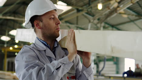 Close-up-view-of-caucasian-carpenter-wearing-helmet-and-carrying-a-big-long-planch-in-a-wooden-factory