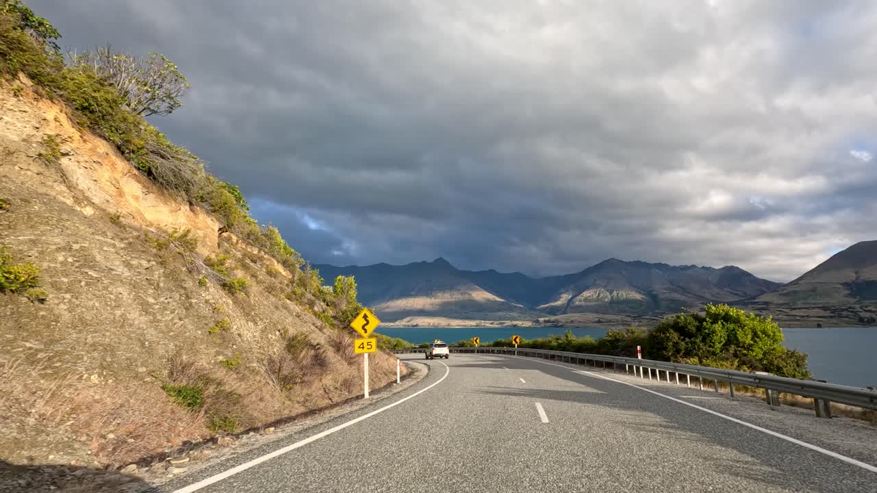 Vehicle travels winding lakeside road, mountain views, dynamic movement, natural daylight, dramatic clouds