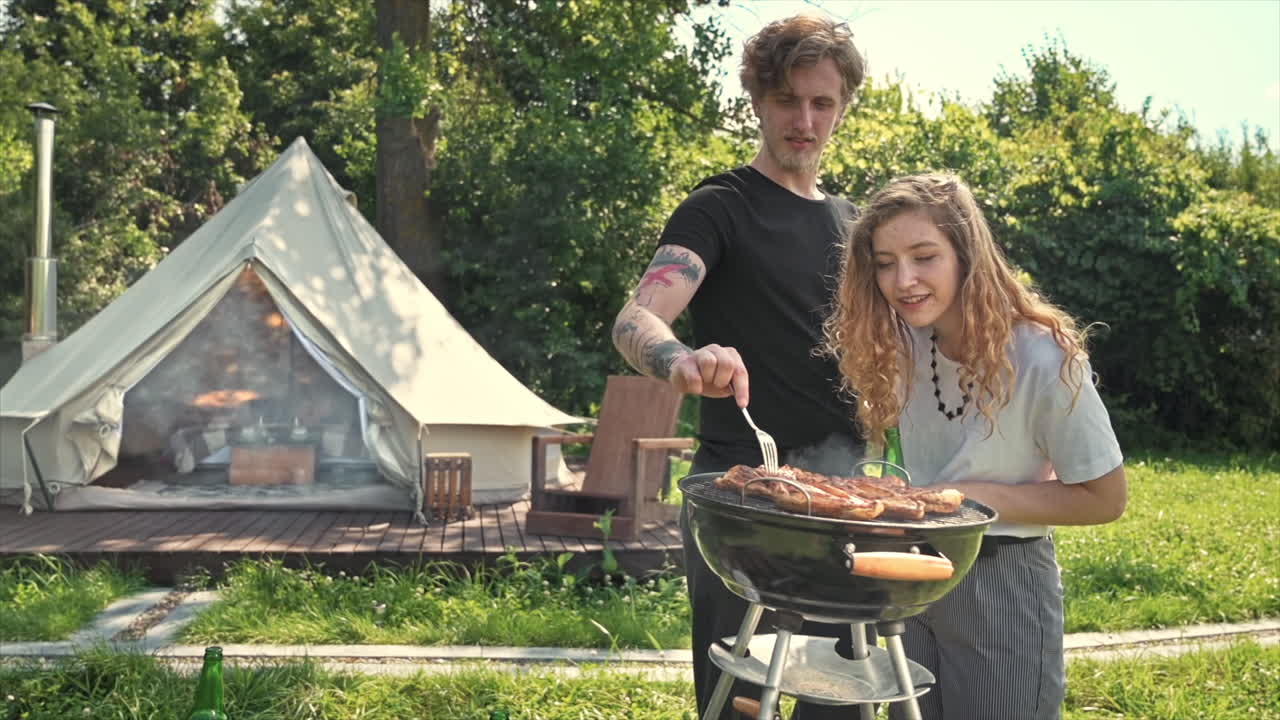 Young couple frying meat on the grill with beer. Greenery around. Glamping tent on the background. Slow motion