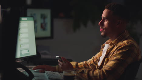 Businessman-Working-Late-In-Office-With-Face-Illuminated-By-Computer-Screen-Using-Mobile-Phone