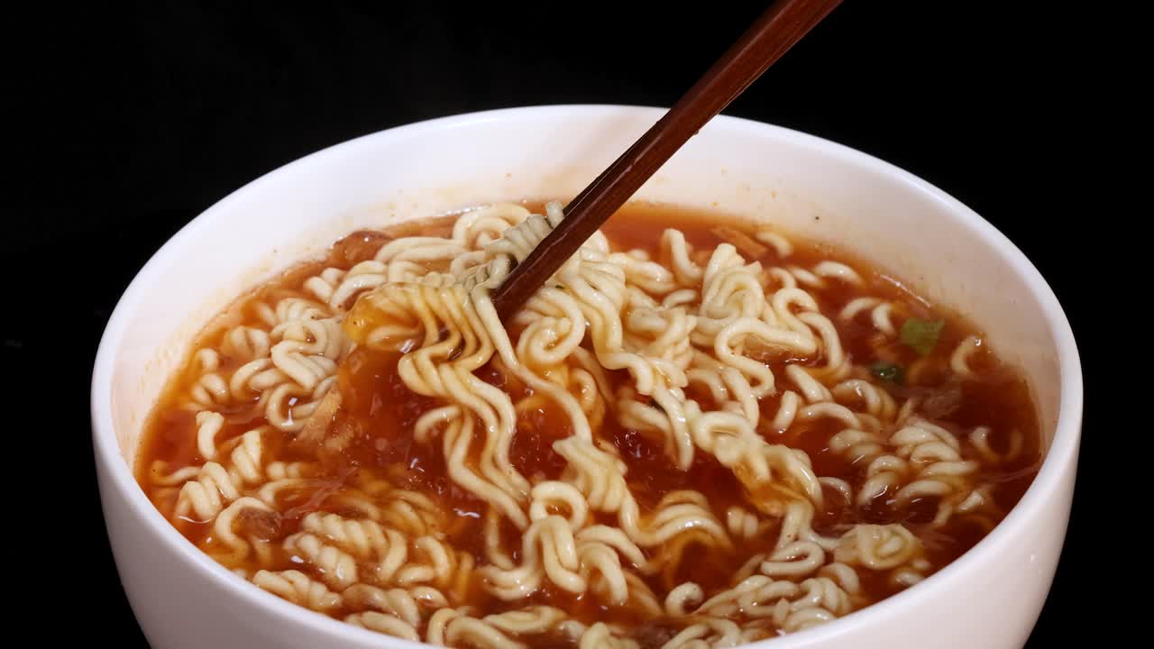 A pair of chopsticks stirs steaming instant noodles in a white bowl filled with broth, under bright studio lighting against a black background. Camera remains steady