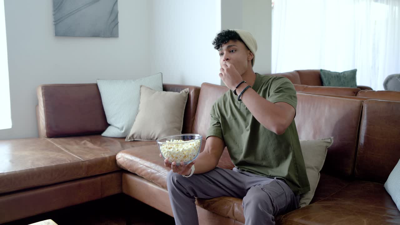 Uomo afroamericano seduto a casa, che guarda fuori campo, con una ciotola di popcorn, mangiando e sorseggiando un bicchiere.