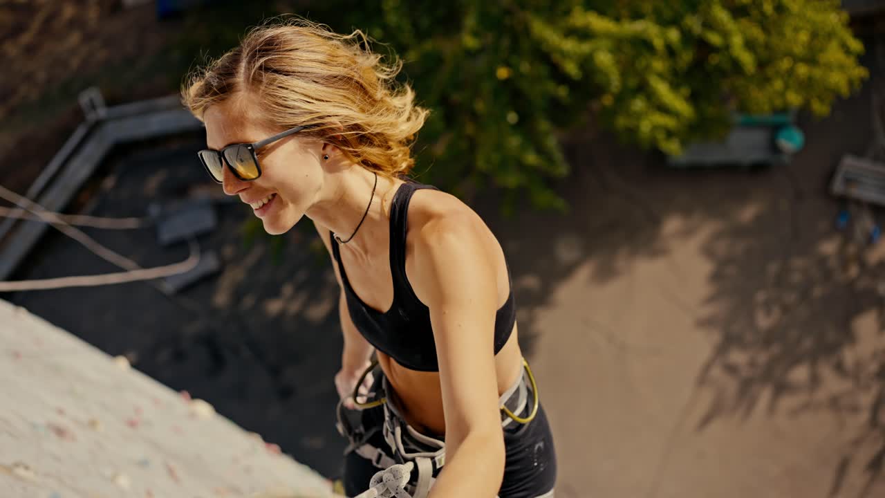 Portrait of a happy blonde girl in a black sports summer uniform and in sunglasses who climbed up a white climbing wall and with multi-colored ledges and poses at a height using a belay in the summer