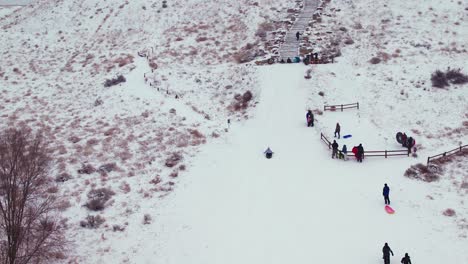 Toma-Aérea-De-Un-Niño-Deslizándose-Por-Una-Empinada-Colina-Cubierta-De-Nieve