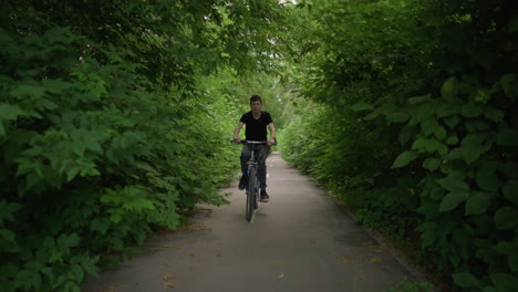 young boy in black top and grey trousers riding a bicycle along a paved path with white markings, surrounded by dense greenery, the ground is littered with leaves