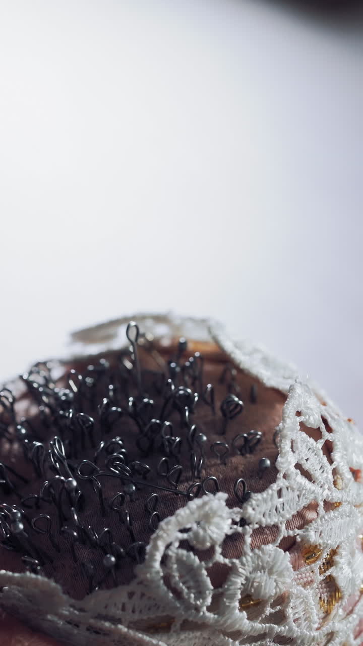 Female hand with beautiful nails taking pins from the white small pincushion. A lot of sewing pins in a sponge for dressmaker on the wooden table. Close-up. Vertical video