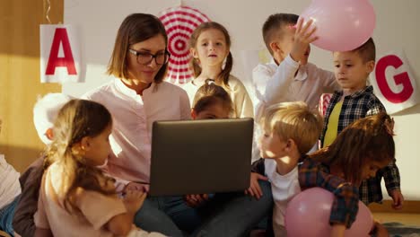 A-female-teacher-with-a-bob-hairstyle,-glasses,-a-fibrous-shirt,-and-a-laptop-are-teaching-a-lesson-for-preschool-children-in-a-special-club-for-preparing-children-for-school