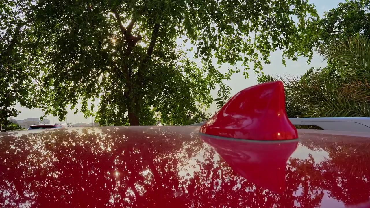 Close-up do teto de um carro vermelho e antena barbatana de tubarão com árvores verdes exuberantes ao fundo. Um brilho solar cinematográfico atravessa as folhas durante o dia claro em um ambiente externo