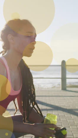 Animation-of-yellow-spots-over-biracial-woman-drinking-water-on-promenade