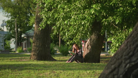 dama sentada al aire libre, bajo un árbol en un campo de hierba, leyendo un libro con la cabeza inclinada ligeramente bajo la cálida luz del sol, las hojas de los árboles se balancean suavemente en la brisa, con el fondo con un edificio residencial